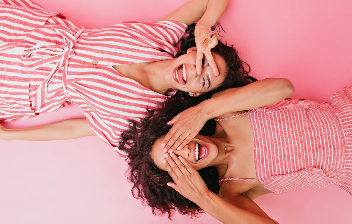 Duas mulheres de vestidos às riscas cor-de-rosa deitadas sobre fundo rosa, a rir e a tapar os olhos com as mãos