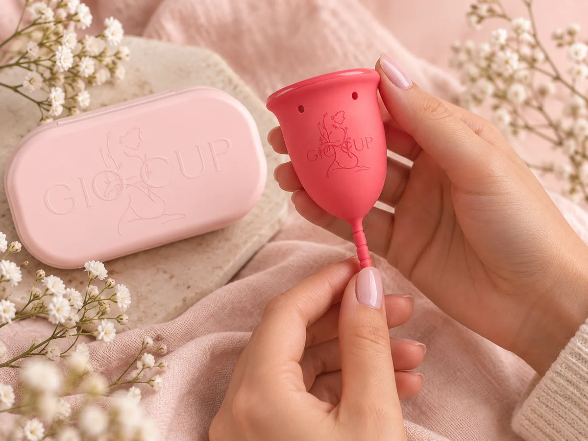 Mãos a segurar um copo menstrual GIOCUP® Rose ao lado do estojo GIOCUP®, entre flores brancas