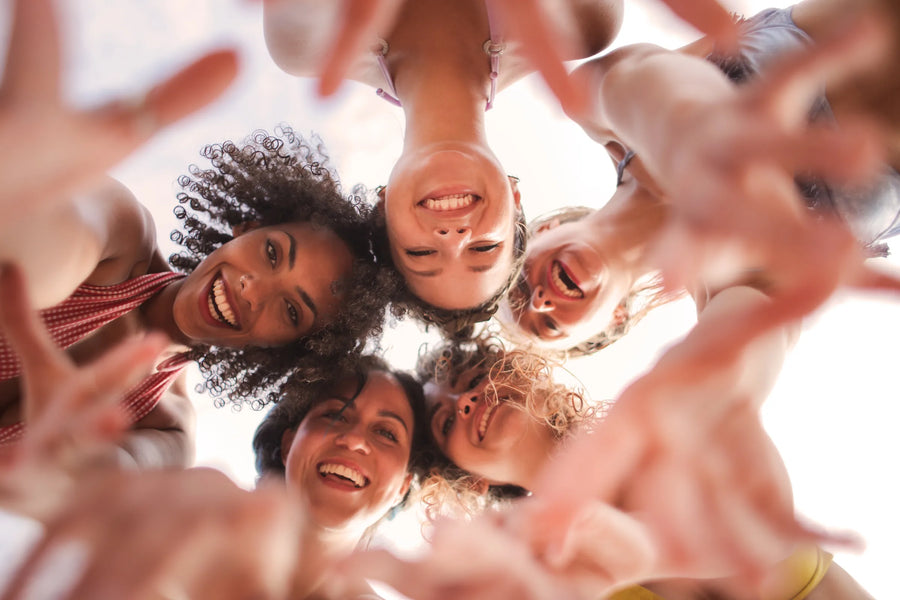 Cinco mulheres em roda, vistas de baixo, a rir com as cabeças juntas e as mãos estendidas